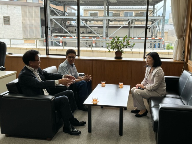 窓から外の景色が見える応接室で、テーブルを挟んで黒いソファに座ったスーツ姿の市長が身振り手振りを交えて話し、その奥には白いシャツ姿の男性、向かいには牧島かれん衆議院議員が座っている視察の様子の写真