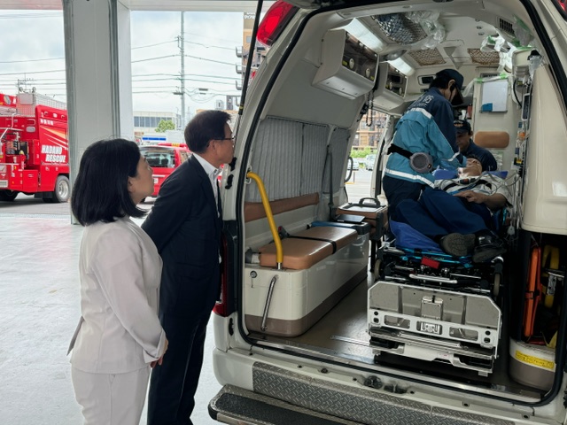 救急車の後部から、救急隊員がストレッチャーに横たわる人の処置している訓練の様子を、スーツ姿の市長と牧島かれん衆議院議員が見学している様子の写真