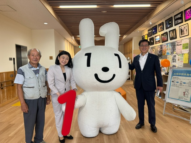 施設内の展示コーナーで、赤い数字の1を持った白いウサギのキャラクター「マイナちゃん」と、グレーのベストを着た男性、牧島かれん衆議院議員、市長が並び撮影した記念写真