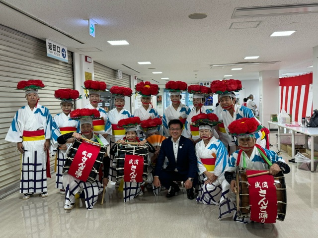 広い廊下で、スーツを着た市長を中央にして、赤い大きな花を飾った笠をかぶり、白い着物を着た男女が多数集まり、数名は太鼓を抱えて撮影した集合写真