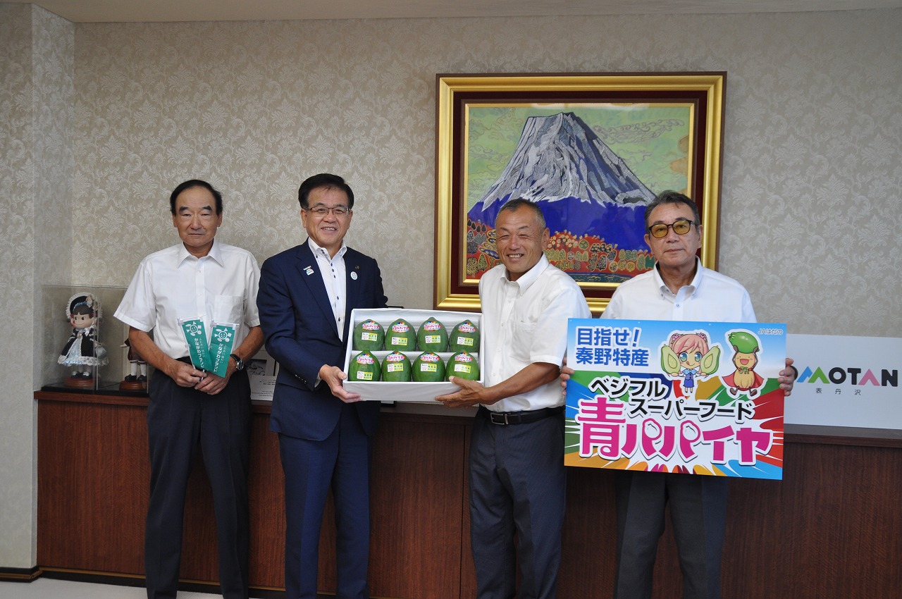 壁に富士山の絵画が飾られた室内で、左から小さな緑ののぼりを2つ持った白いシャツの男性、8個の青パパイヤが入った箱を両側から持った市長と白いシャツの男性、青パパイヤの宣伝パネルを持った男性が並んで立って写した記念写真