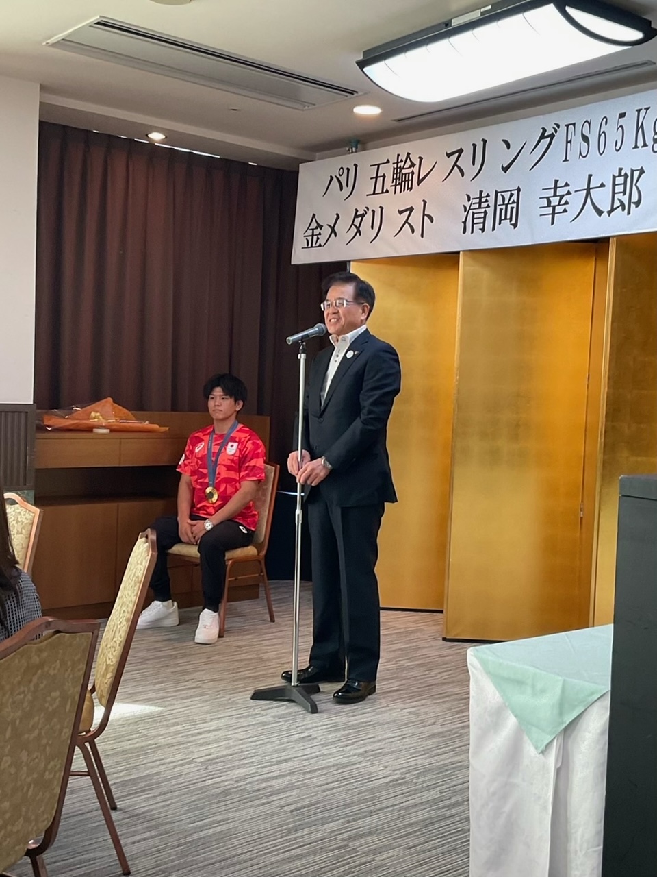 「パリオリンピック レスリングFS65kg 金メダリスト 清岡幸大郎」と書かれた横断幕が掲げられた金屏風の前で、赤いTシャツと黒いズボンを着用し金メダルを首から下げた清岡選手が椅子に座っている横で、黒いスーツ姿の市長がマイクでスピーチをしている写真