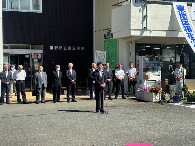秦野市立東公民館前の広場で、数名のスーツ姿や白いシャツ姿の男性たちが立って整列している前で、スーツを着た市長がマイクで話をしている写真