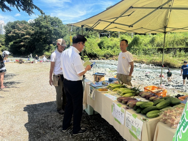晴天の河原で、白いワイシャツを着た市長が、新鮮なトウモロコシやナス、キュウリなどの夏野菜を販売しているテントの前に立ち、白いTシャツ姿の男性と話をしている様子を写した写真