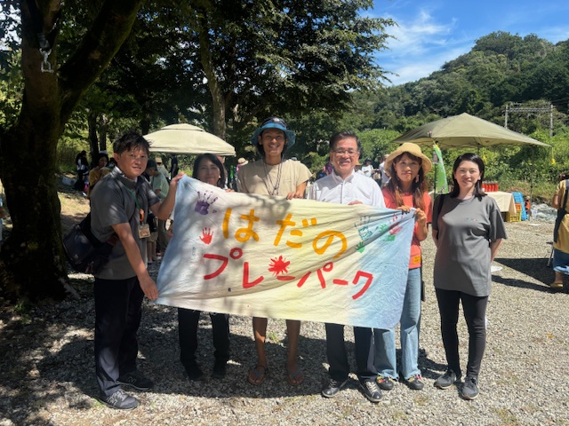 川沿いの屋外イベント会場で、日差しを浴びて木陰の下に立つ5人の男女と市長が、「はだのプレイパーク」と書かれたカラフルな横断幕を広げて持ち、笑顔で立っている記念写真