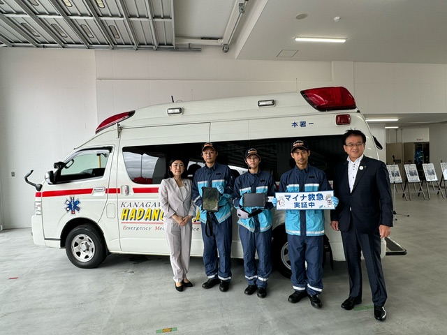 車庫に駐車した救急車の前で、牧島かれん衆議院議員、紺色の制服を着た男性救急隊員3名、市長の計5名が並んで写した記念写真