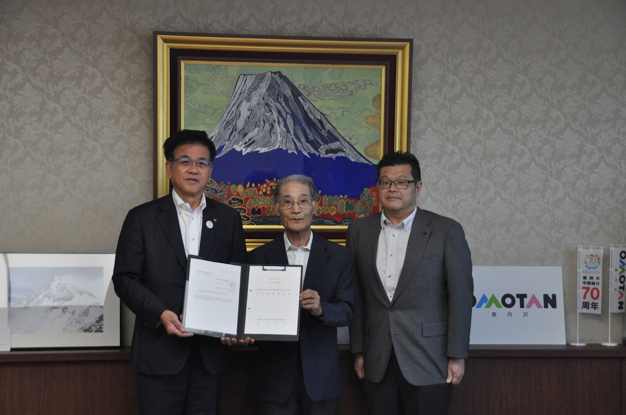 背景の壁に飾られた富士山の絵画の前で、黒いスーツの男性が中央に立ち、左側のスーツ姿の市長と意見書を持ち、右側にはグレーのスーツ姿の男性が立っている記念写真