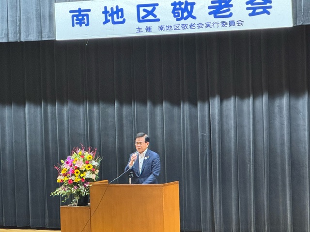 「南地区敬老会」と書かれた横断幕が掲げられているホールの舞台上で、紺色のスーツを着た市長が木目調の演壇でマイクを使いスピーチをしているイベントの様子の写真