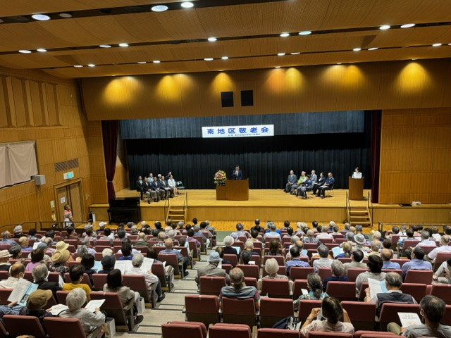 「南地区敬老会」と書かれた横断幕が掲げられたステージの上で市長が演台に立ち、その両脇には複数のスーツ姿の男性が並んで着席し、会場には多数の聴衆が座っている、会場全体の様子を客席側から写した写真