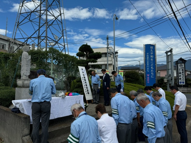 道路脇の敷地内で、台座の上に立つ石仏の前に、白い布をかけた献花台が設けられ、水色のシャツに緑の腕章をつけた複数の男性や、ワイシャツ姿の男性らが集まり、石仏に頭を下げて黙祷を捧げている様子の写真