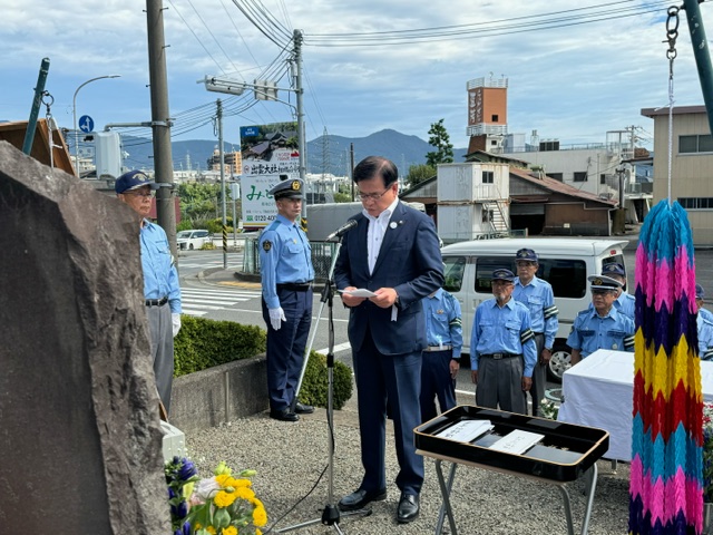 交差点を背景に、紺色のスーツを着た市長が花が添えられ、千羽鶴が飾座られた石碑の前に設置されたマイクに向かい、原稿を読み上げており、その後ろには水色のシャツを着た警察官や交通安全指導員の男性たちが姿勢を正して立っている写真