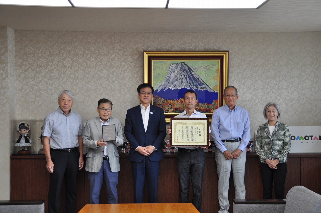 富士山の絵画が飾られた壁の前で、中央に立った紺色のスーツの市長の左右に、男女計5名が並んでおり、左から2番目の男性は記念品の盾、右から3番目の男性は額に入った賞状を胸の前に掲げている記念写真