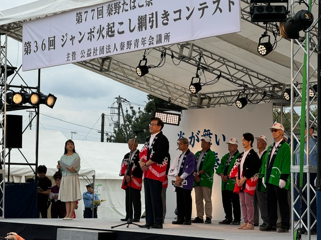 野外の特設ステージ上で、赤と黒の配色の法被を着た市長がマイクで話し、左奥に進行役の女性、市長の後ろに緑や黒などの法被を着た複数の関係者の男女が並んで立っている写真