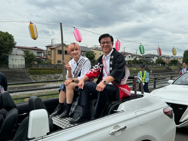 屋根を開けた白いオープンカーの後部座席に、ピンク色のショートカットの女性と、黒と赤の柄の法被を着た市長の2人が座り、色とりどりの提灯が飾られた道をパレードしている様子の写真