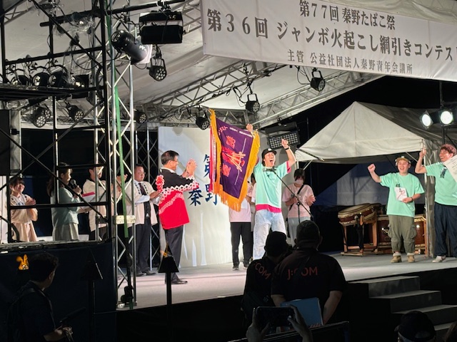 夜間の特設ステージ上で、黒と赤の配色の法被を着た市長が拍手する横で、淡い緑色のTシャツを着た男性が、紫色の旗を掲げ喜びを表現し、右側では同じ薄い緑色のTシャツを着た男性2人がガッツポーズをしている、表彰式の様子の写真