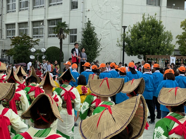 野外の広場で、編み笠を被り白地に緑色の柄が入った着物に赤の襷をつけた女性の団体と、青い法被を着てオレンジ色の帽子を被った団体が整列して立っており、前方の台に上がった市長の話を聞いている様子を後方から撮影した写真