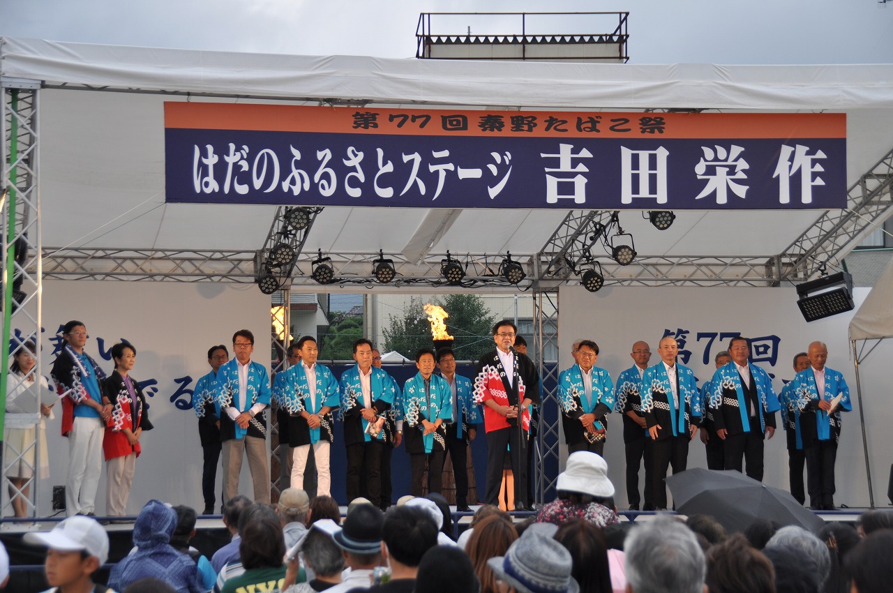 「はだのふるさとステージ 吉田栄作」と書かれた横断幕が設置された屋外特設ステージ上で、黒と赤の配色の法被を着た市長がステージ中央に立ち、その後ろに水色と黒の配色の法被を着た関係者がたくさん並んで立っている写真