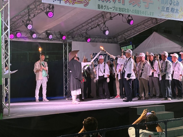 夜間の屋外特設ステージ上で、黒い着物に編笠を被った人物が中央に立ち、向かいあって、列になって立った白い法被と帽子を身に着けたたくさんの人々の先頭の男性に、たいまつの火を受け渡すような動作をしている、ステージの催し物の様子の写真