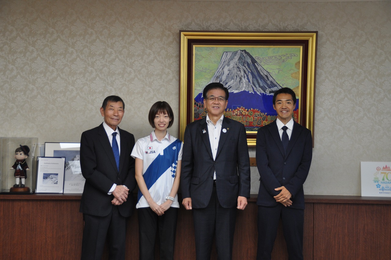 壁に富士山の絵画が飾られた部屋で、左からスーツ姿の男性、青いタスキをかけた池神悠希さん、スーツ姿の市長、スーツ姿の若い男性の計4人が並んだ記念写真