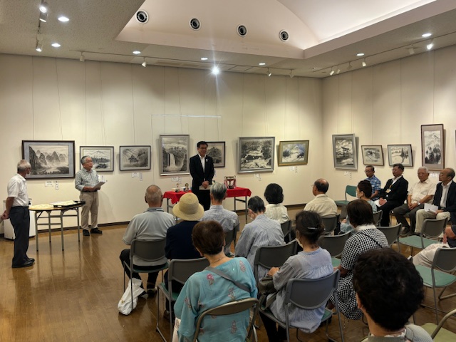正面や右側の壁に水墨画が多数展示された会場で、並んだ椅子に着席したたくさんの出席者に向かって、前方に立った市長が話をしている様子の写真