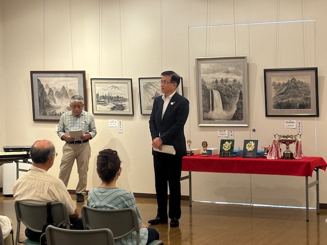 山や滝などが描かれた複数の水墨画が飾られた壁の前で、白いチェック柄のシャツを着た男性が手に持った原稿を読み上げており、中央に置かれた、赤い布が敷かれ、複数のトロフィーや盾が並んだ長机の前に立った市長と、着席した出席者たちが男性の話を聞いている様子の写真