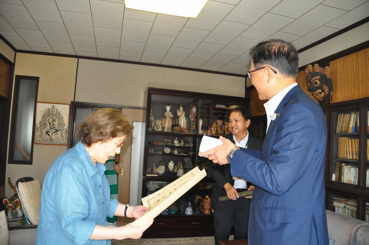 骨董品や書籍が並んだ室内で、青いシャツを着た年配の女性が、藍色のスーツを着た市長から賞状を手渡されている様子を後方でスーツ姿の別の男性が笑顔で見守っている、贈呈式の様子を捉えた写真