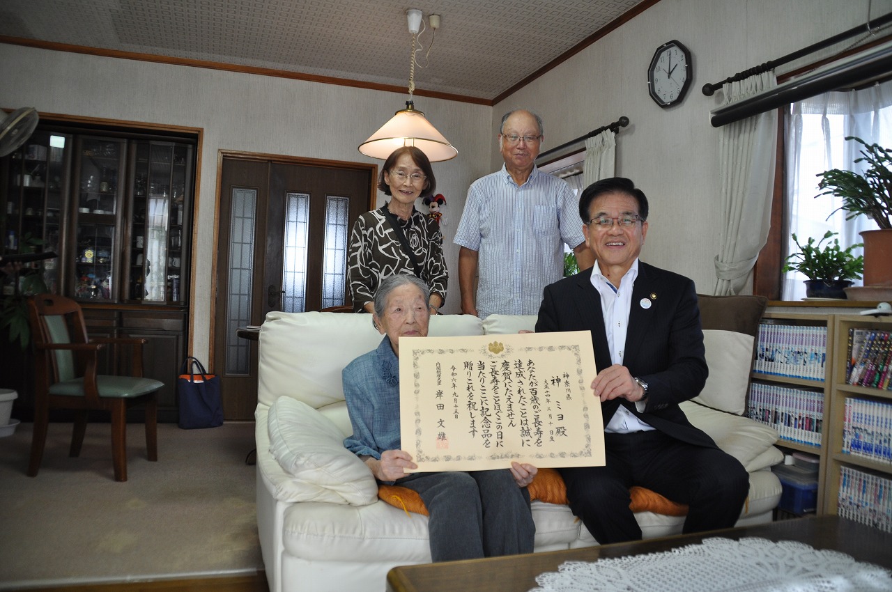 リビングルームで、白いソファに座った高齢の女性と黒いスーツ姿の市長が賞状を広げて持ち、その後ろに年配の男女2人が立ってカメラに向かって微笑んでいる写真