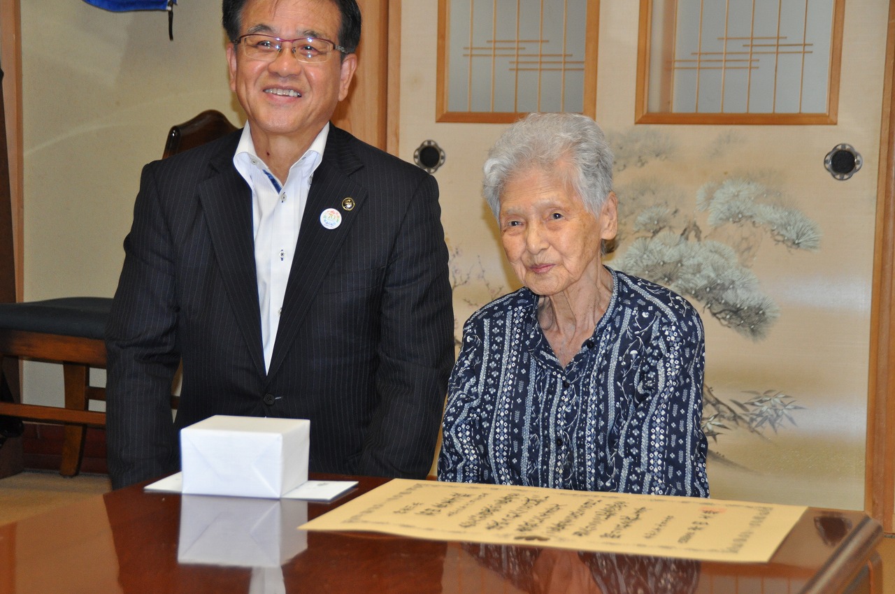 白い箱と賞状が置かれた座卓を前に、縞模様の服を着た高齢の女性と、紺色のスーツを着た市長が座り、笑顔を見せている記念写真