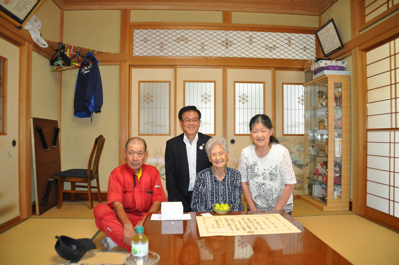 和室で座卓を前に、赤いつなぎの年配男性、黒いスーツの市長、縞模様の服の高齢女性、白い服の年配女性の4人が笑顔で並んで撮影した記念写真
