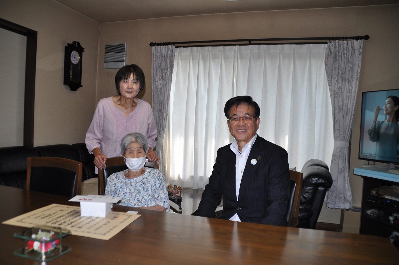白い箱と賞状が置かれた木製のダイニングテーブルを前に、黒いスーツ姿の市長とマスクをした高齢の女性が座り、後ろに立った薄いピンク色のブラウスの女性と一緒に撮影した記念写真