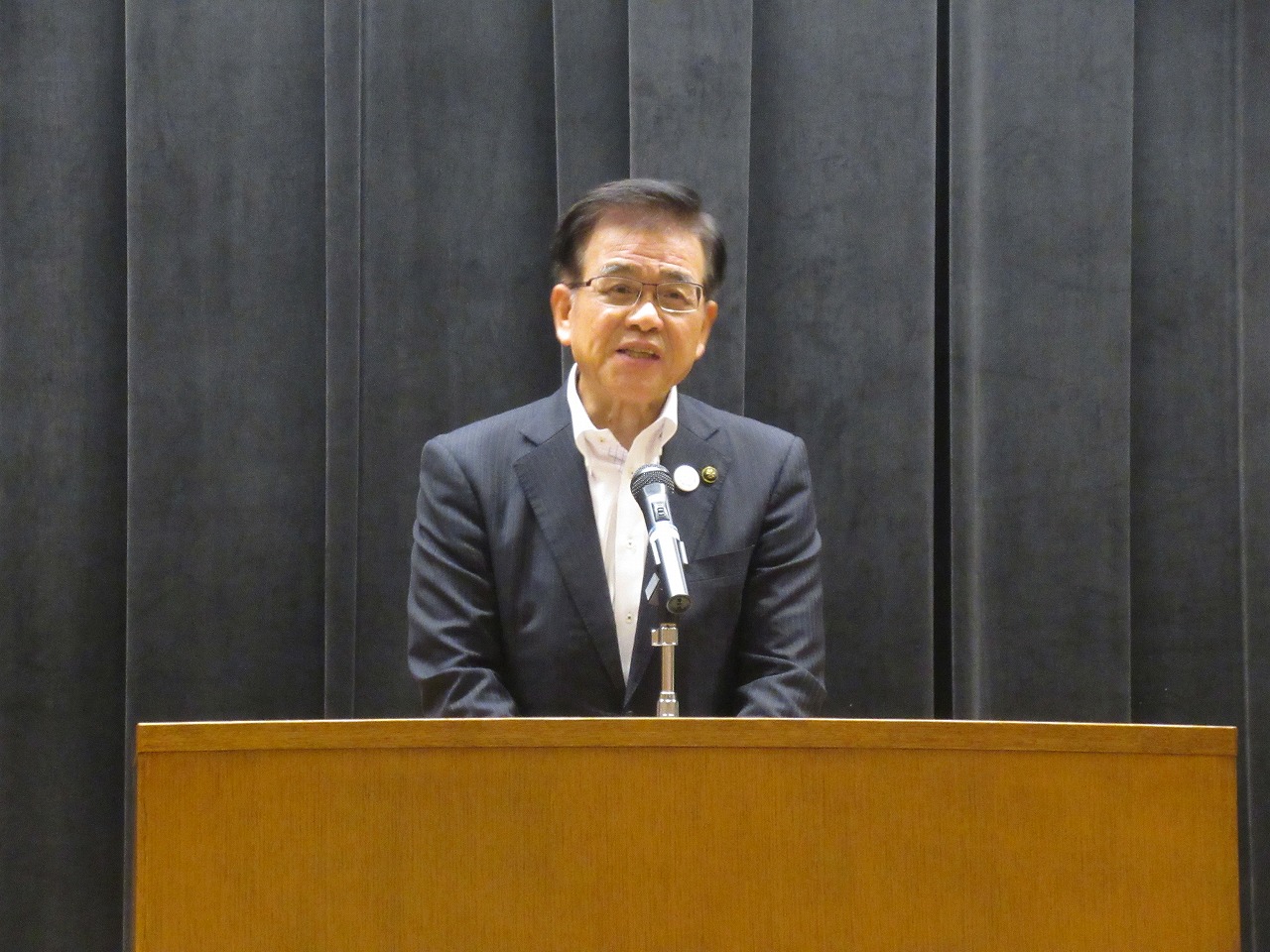 黒いカーテンの前で、スーツ姿の市長が演台に立ち、マイクに向かって話している写真