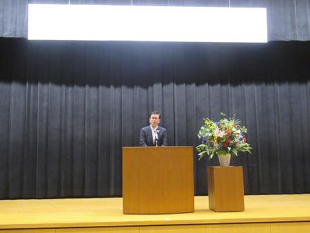 黒いカーテンのステージ上で、右隣に色鮮やかな花が飾られた演壇の前に立つスーツ姿の市長を撮影した写真