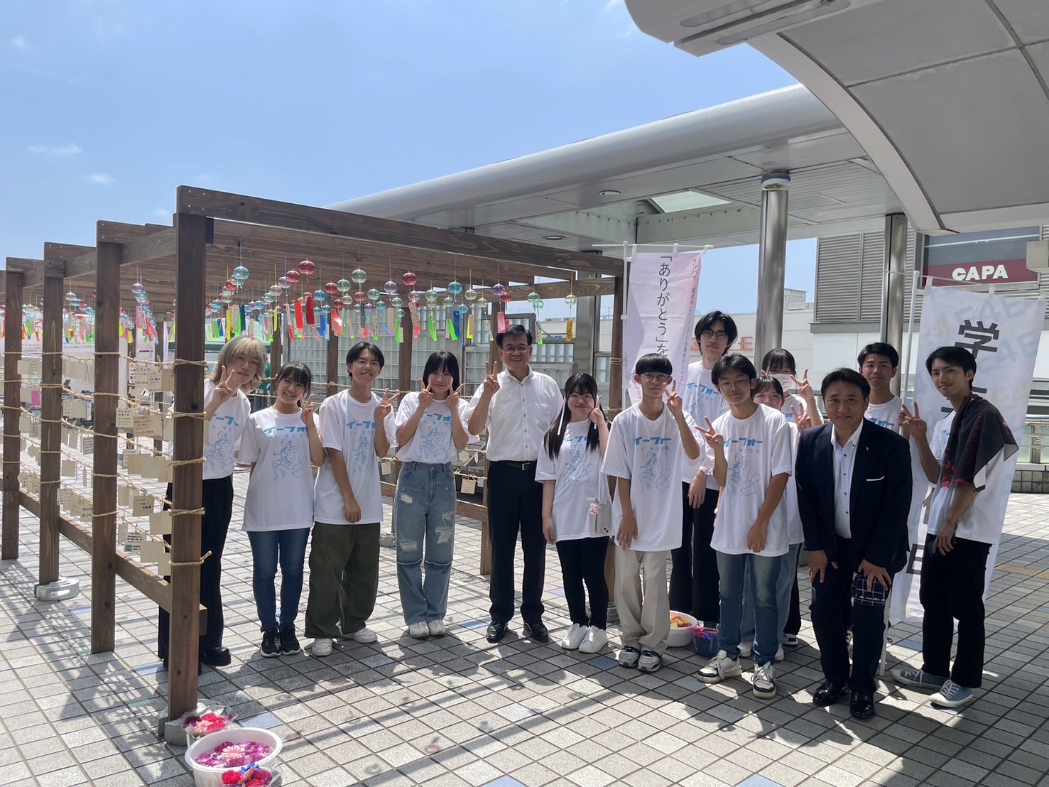 青空の下、風鈴が飾られた場所で、お揃いの白いTシャツを着た学生たちと、市長と関係者が集まってピースサインで記念撮影をしている写真