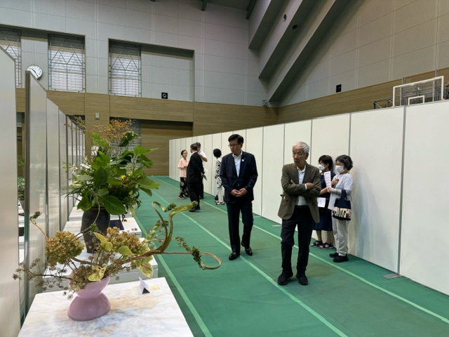 体育館のような広い会場で、緑色のトラック上を紺色のスーツを着た市長と茶色のジャケットの年配男性が歩きながら、仕切り板に沿って並べられた生け花の展示作品を鑑賞している、文化展の様子の写真