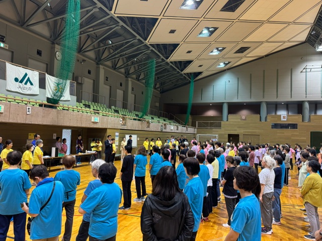 体育館で多くの参加者が整列しており、前方でマイクを持ったスーツ姿の男性が話をしている様子の写真
