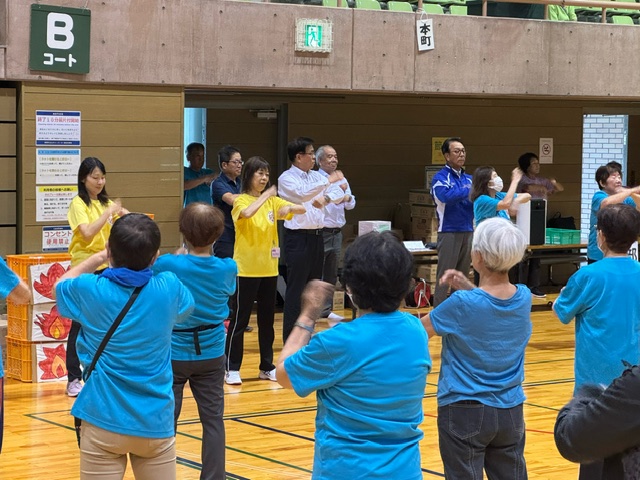 体育館の中で多くの参加者が運動をしており、最前列には黄色のTシャツを着た数人が見本を見せている、体操イベントの様子の写真