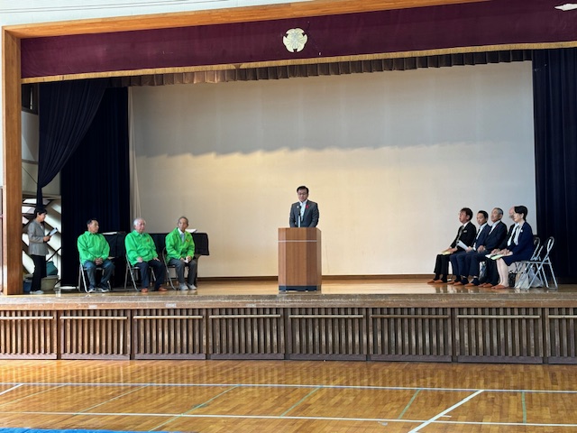 体育館のステージ上で、演台に立つ市長がスピーチをしており、その両側に数人の関係者が着席している式典風景の写真