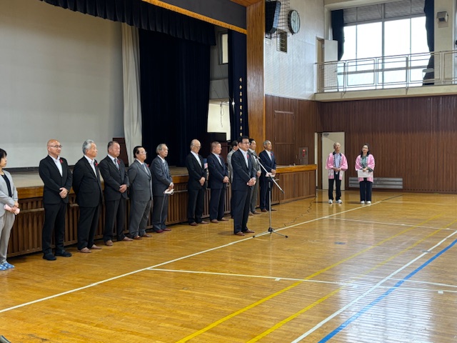 体育館のステージ下で、多くのスーツ姿の男女が整列しており、その前でマイクに向かって話をしている市長を捉えた式典風景の写真