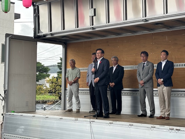 トラックの荷台を改装した舞台で、市長がマイクを持って挨拶をしており、他の数人の男性が両脇に立っている屋外イベントの写真