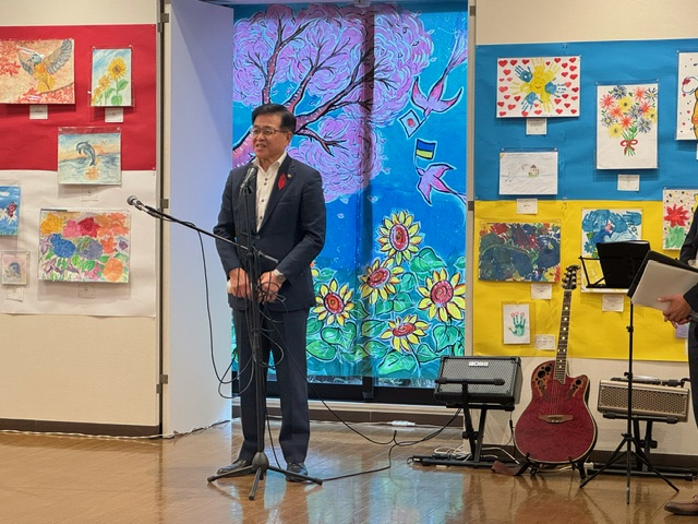 様々な子供たちの絵が壁に展示された会場で、市長がマイクの前に立って挨拶しており、近くにはギターとアンプが置かれているイベント風景の写真