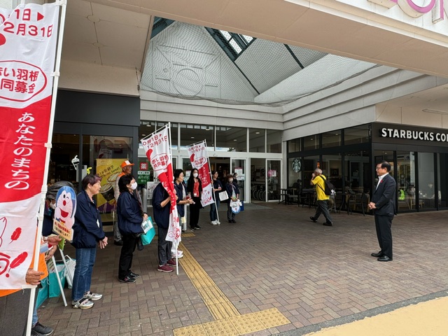 「赤い羽根共同募金」と書かれたのぼり旗と募金箱を持った人々が、向かい合わせになった市長の話を聞いている募金活動の様子の写真