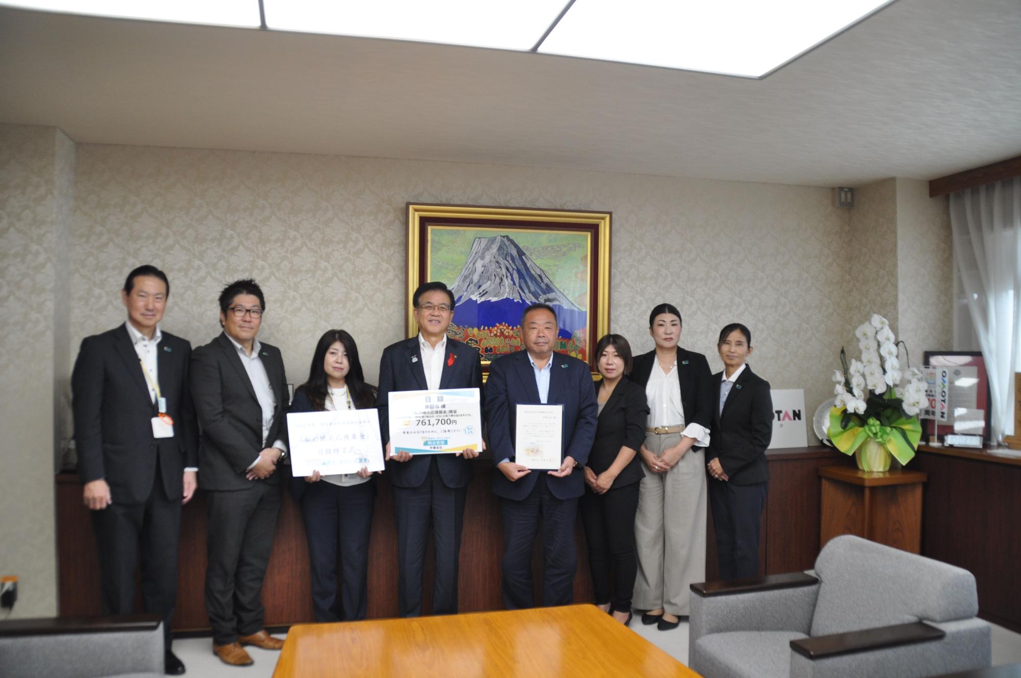 富士山の絵が飾られた部屋で、スーツを着用した市長を含めた8人の男女が並んでおり、中央の3名が大きなボードや賞状のようなものを持って記念撮影している写真