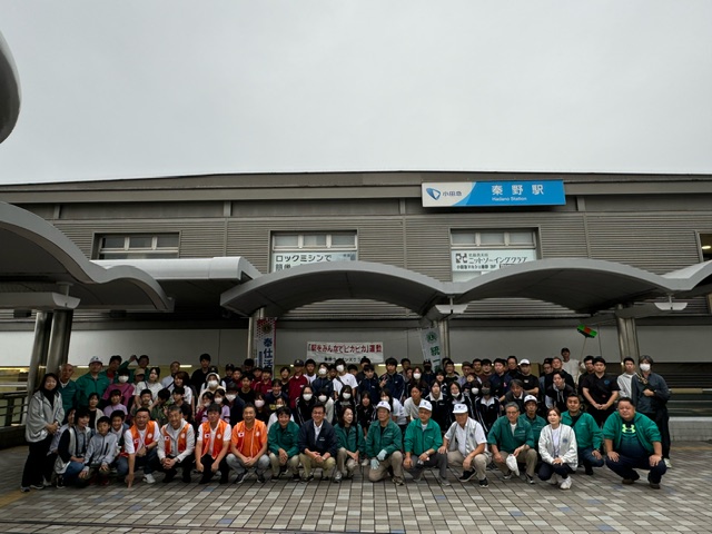 小田急線の秦野駅の駅舎を背景に、多くの人が整列してカメラに向かってポーズを取っている集合写真