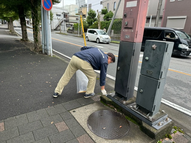 歩道で紺色の上着とベージュのズボンを履いた市長と思われる男性が、透明なゴミ袋を片手に、電柱の根元のごみを拾っているピカピカ運動の様子写真