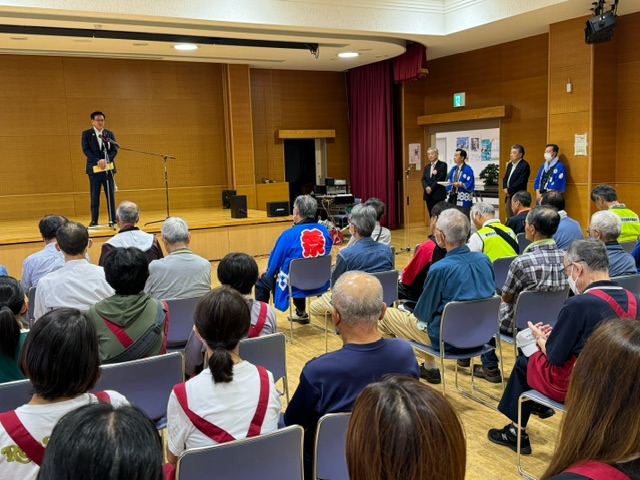 室内イベントの壇上で市長がスピーチをしており、複数の関係者と多くの観客が着席しているまつり会場の写真