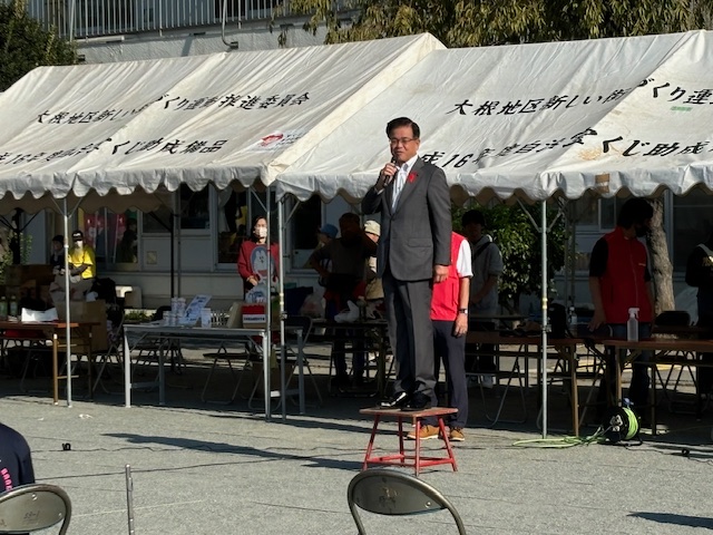 テントが並ぶ屋外会場で、市長が小さな台の上に立って、マイクを使い話しているイベント風景の写真