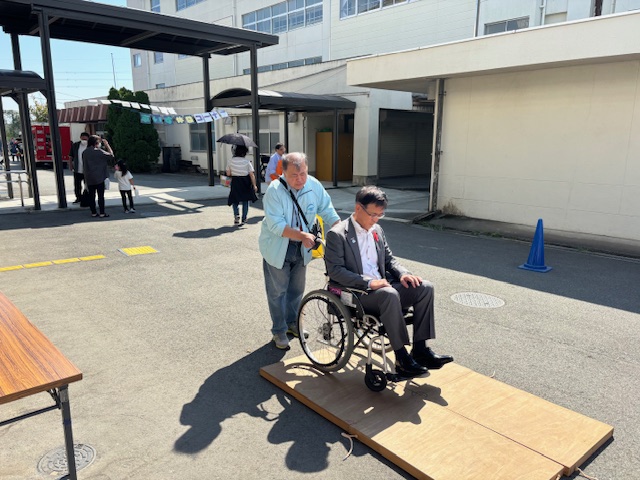 屋外のイベント会場で、スーツ姿の市長が車いすに乗ってスロープを体験しており、後ろから別の男性が支えている写真