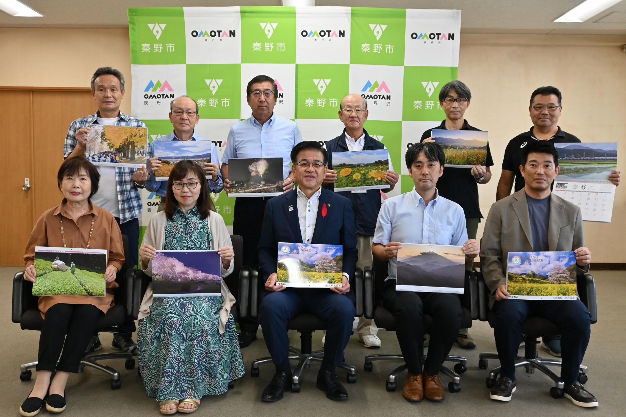 秦野市のバックボードの前で市長を含めた複数の男女が秦野市の風景写真が載ったカレンダーを持ち記念撮影を行っている写真