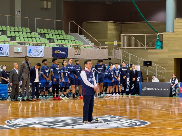 アリーナのコート上で、マイクを持って話すユニフォームを着用した市長の後ろに、紺色のユニフォームを着たバスケットボールチームの選手たちが整列している式典風景の写真