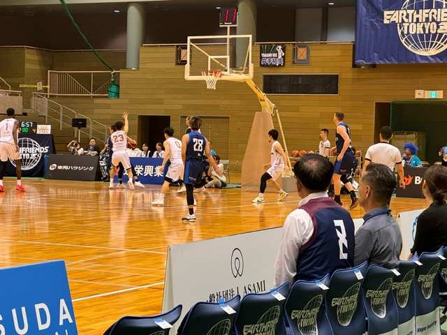 バスケットボールの試合会場で、ユニフォームを着た選手たちがコート上でプレイしており、観客席の椅子には複数人の観客が座っている写真